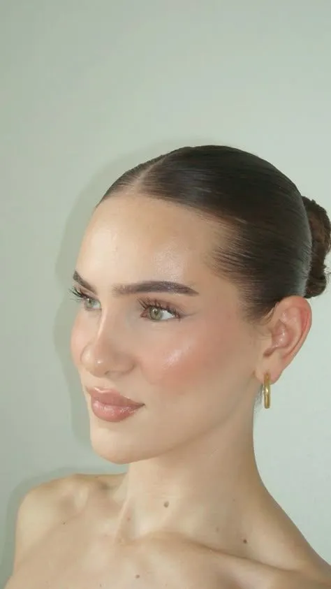Clean Girl aesthetic — Glazed dewy skin, brushed-up brows, small gold hoops, and a slicked-back bun are all signature elements.