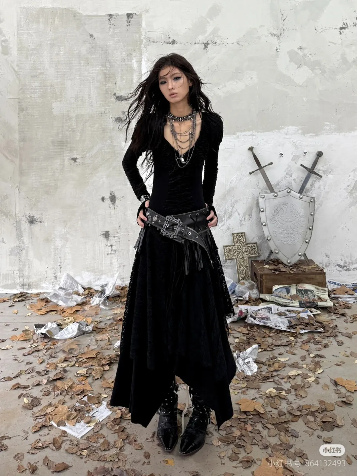 Darksynth aesthetic — The combination of a black lace dress, studded belt, layered necklaces, and the presence of a cross and shield in the background strongly aligns with the occult symbolism, signature garments, and horror-inspired visual cues.