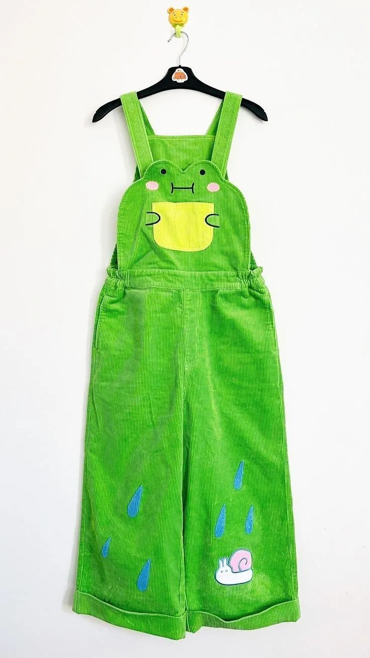 Frogcore aesthetic — The bright green corduroy overalls with a frog face appliqué and rain/snail details directly reference the frog-print everything and mushroom/pond-life motifs central to Frogcore.