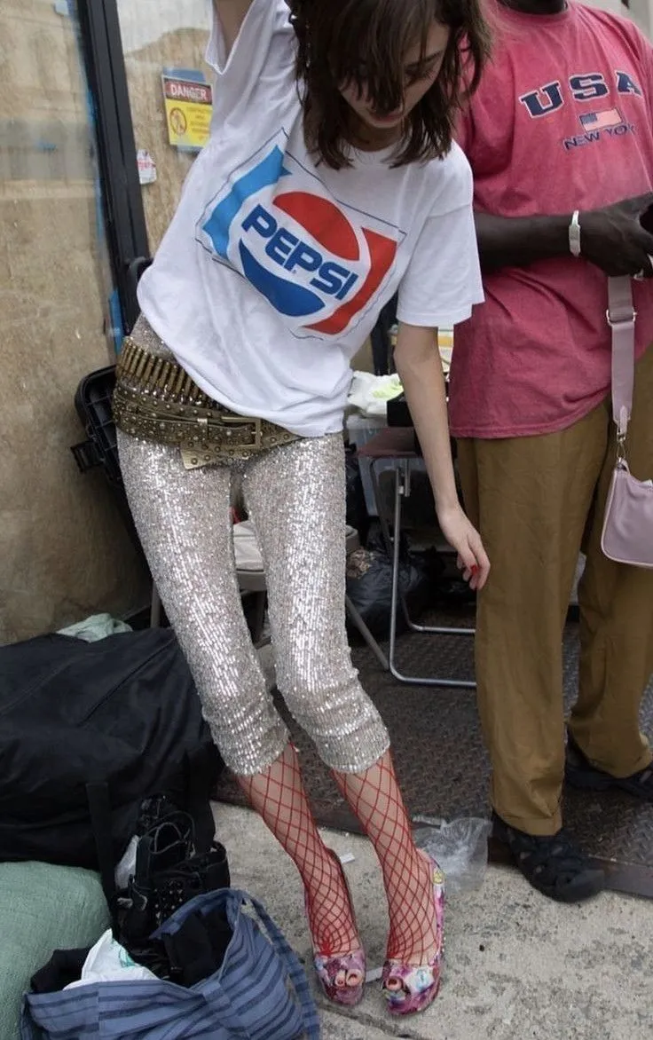 Indie Sleaze aesthetic — Sequined disco pants, ripped fishnets, and a band tee are direct references to the description's signature garments and overall messy high-low style.