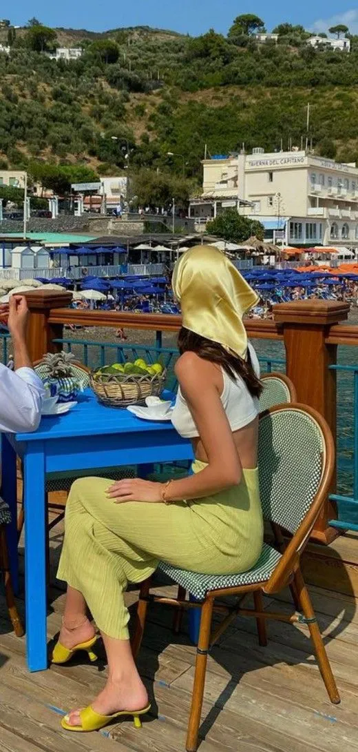 Italian Summer aesthetic — Silk headscarf, linen-like trousers, yellow mules, and the seaside setting perfectly capture the Italian Summer aesthetic.