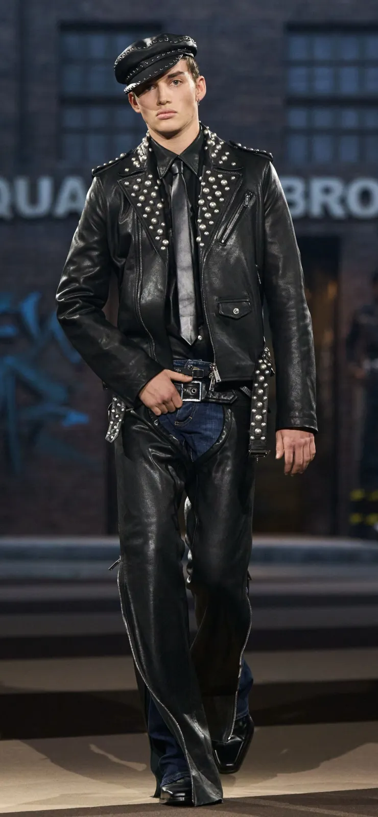 Leather Subculture aesthetic — The combination of a studded leather jacket, chaps over jeans, peaked cap, and tie is a direct and accurate representation of the described visual codes.