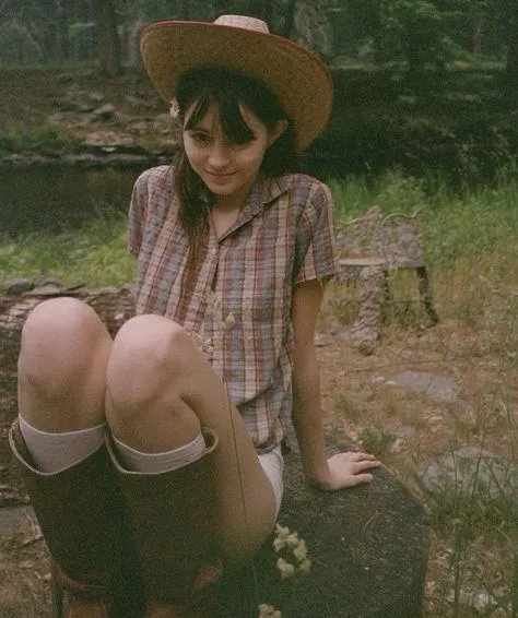 Midwest Gothic aesthetic — The sun-bleached plaid shirt, work boots, straw hat, and outdoor, slightly unkempt setting perfectly capture the described visual signature and mood of Midwest Gothic.