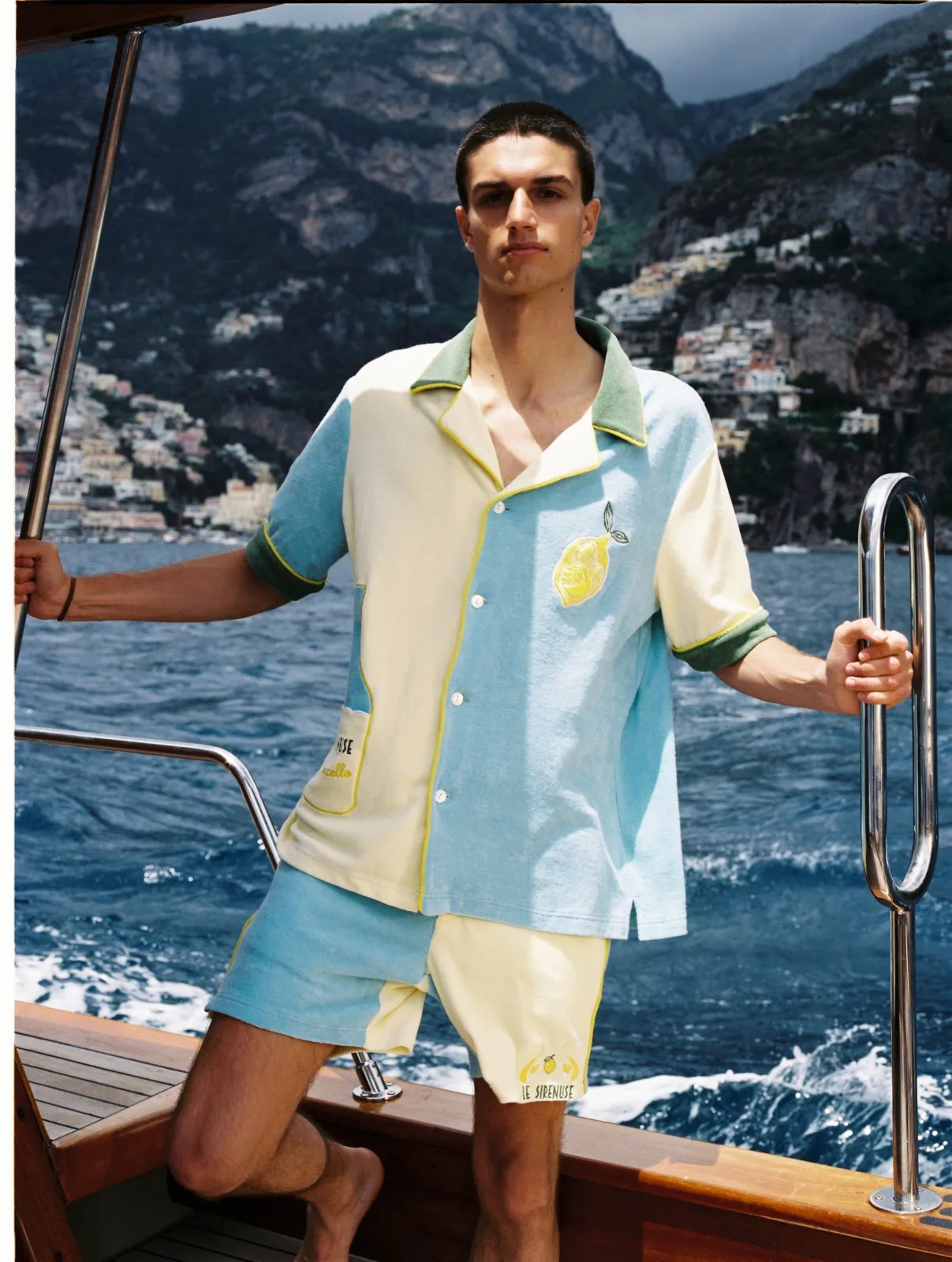 Resortwear aesthetic — The color-blocked shirt and shorts set with a lemon motif, worn on a boat with a coastal backdrop, is a perfect representation of resortwear.