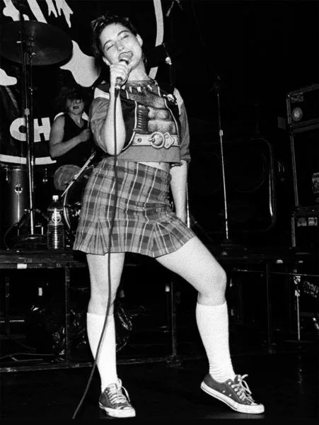 Riot Grrrl aesthetic — Plaid school skirt, knee socks, and a vintage tee worn by a performer capture the essence perfectly.
