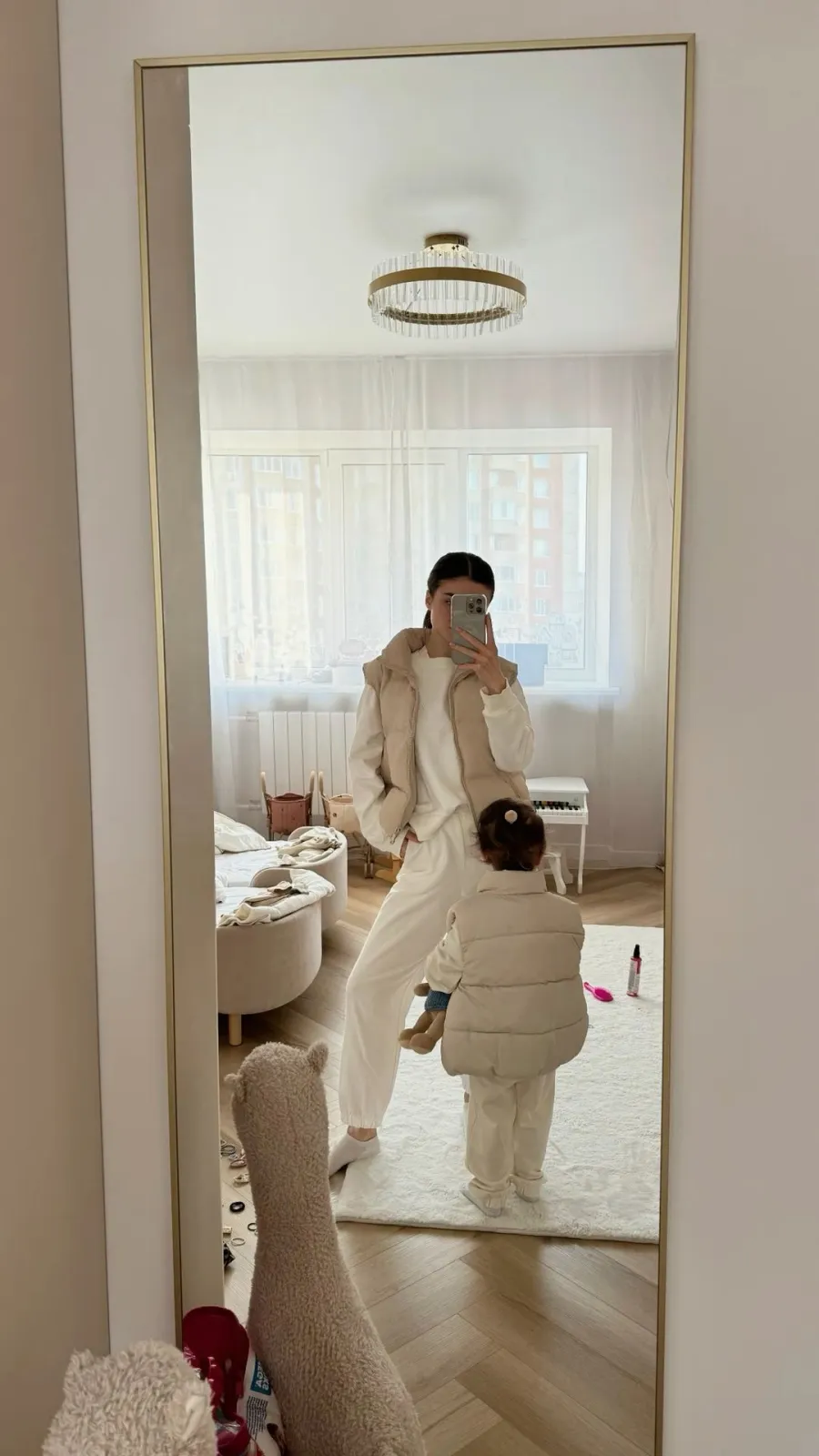 Sad Beige aesthetic — The matching neutral puffer vests for mother and child, along with the neutral-toned nursery background, directly reflect the 'mommy-and-me' and nursery aspects of the Sad Beige aesthetic.
