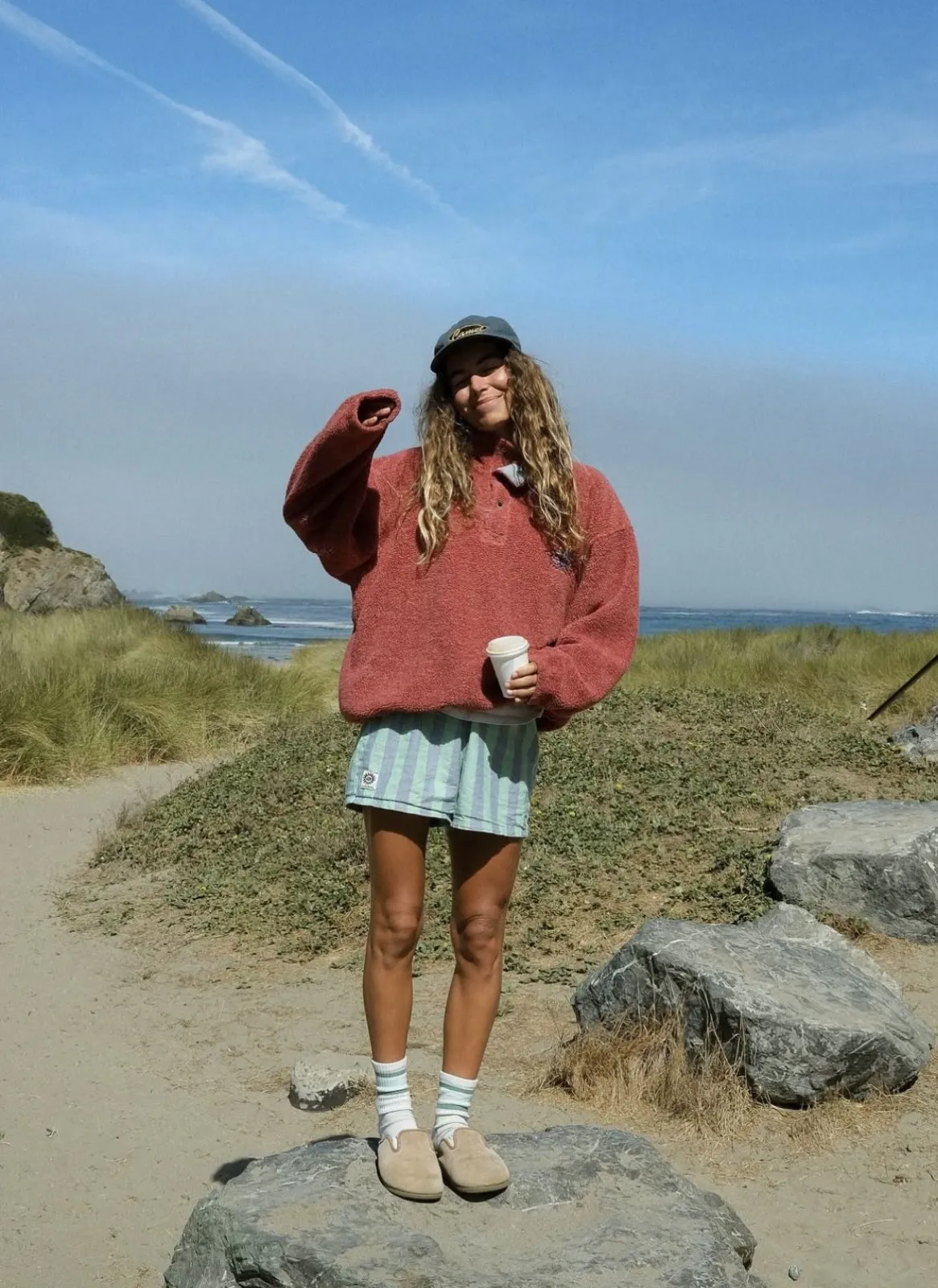 Surfcore aesthetic — The model's sun-bleached, messy hair, fleece, board shorts, and beach setting are quintessential Surfcore.