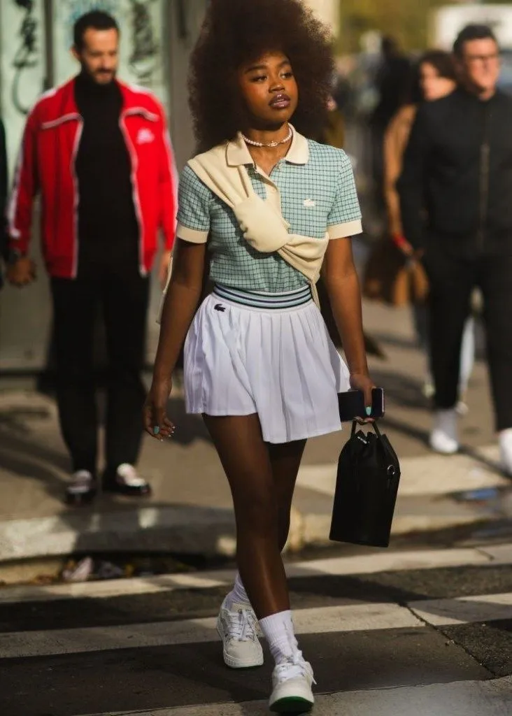 Tenniscore aesthetic — Combines a pleated tennis skirt, polo shirt, and cable-knit sweater draped over the shoulders, hitting multiple signature elements.