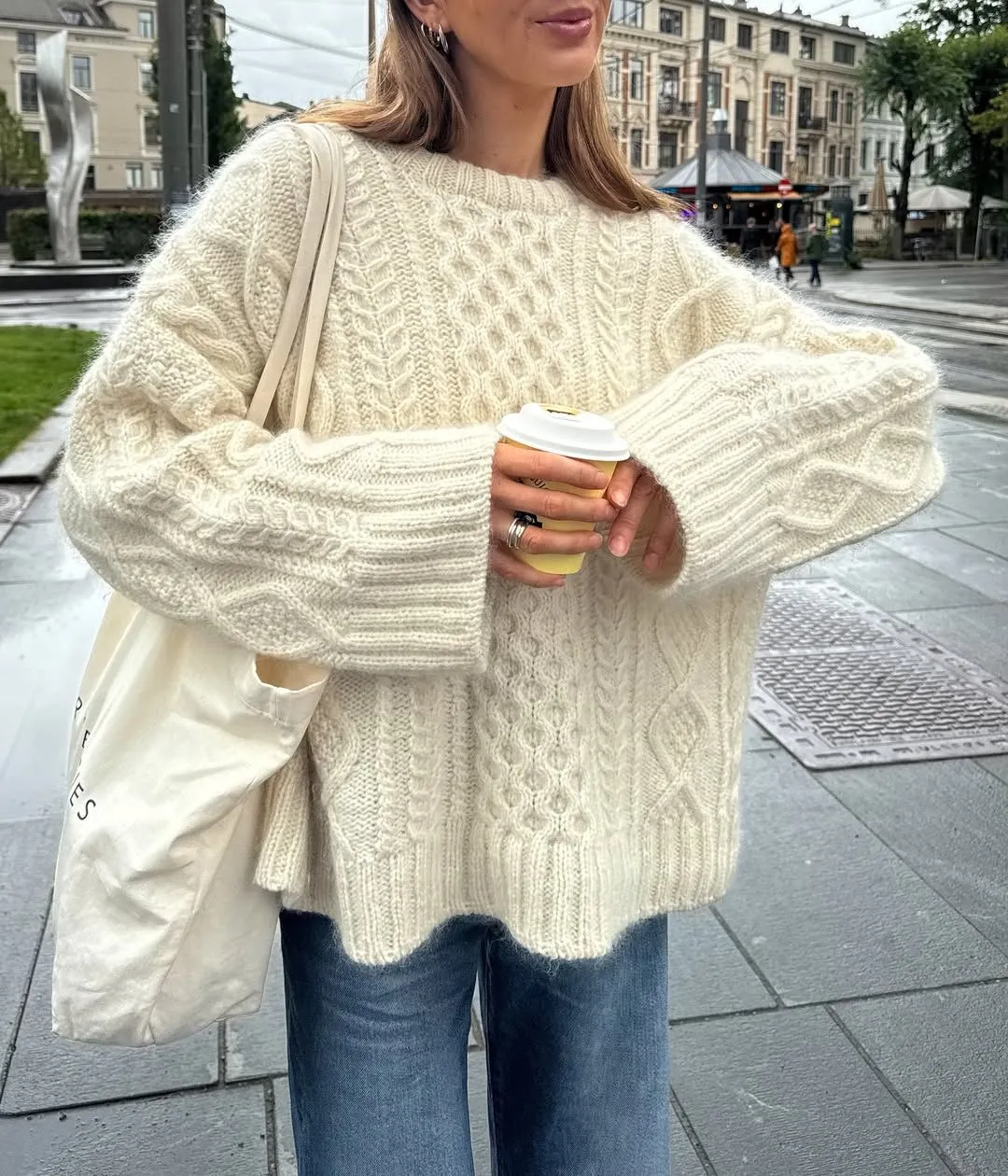 Vanilla Girl aesthetic — Woman holding an oat milk latte in a cream cable knit sweater with a tote bag captures the visual signature and cozy, everyday feel.