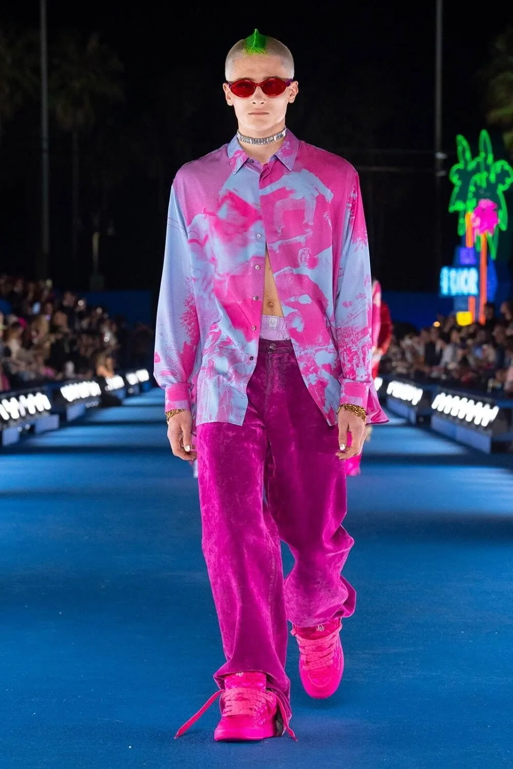 Vaporwave aesthetic — The hot pink and pastel color palette, the oversized camp-collar shirt with a graphic print, and the neon palm trees in the background strongly evoke the Vaporwave aesthetic.
